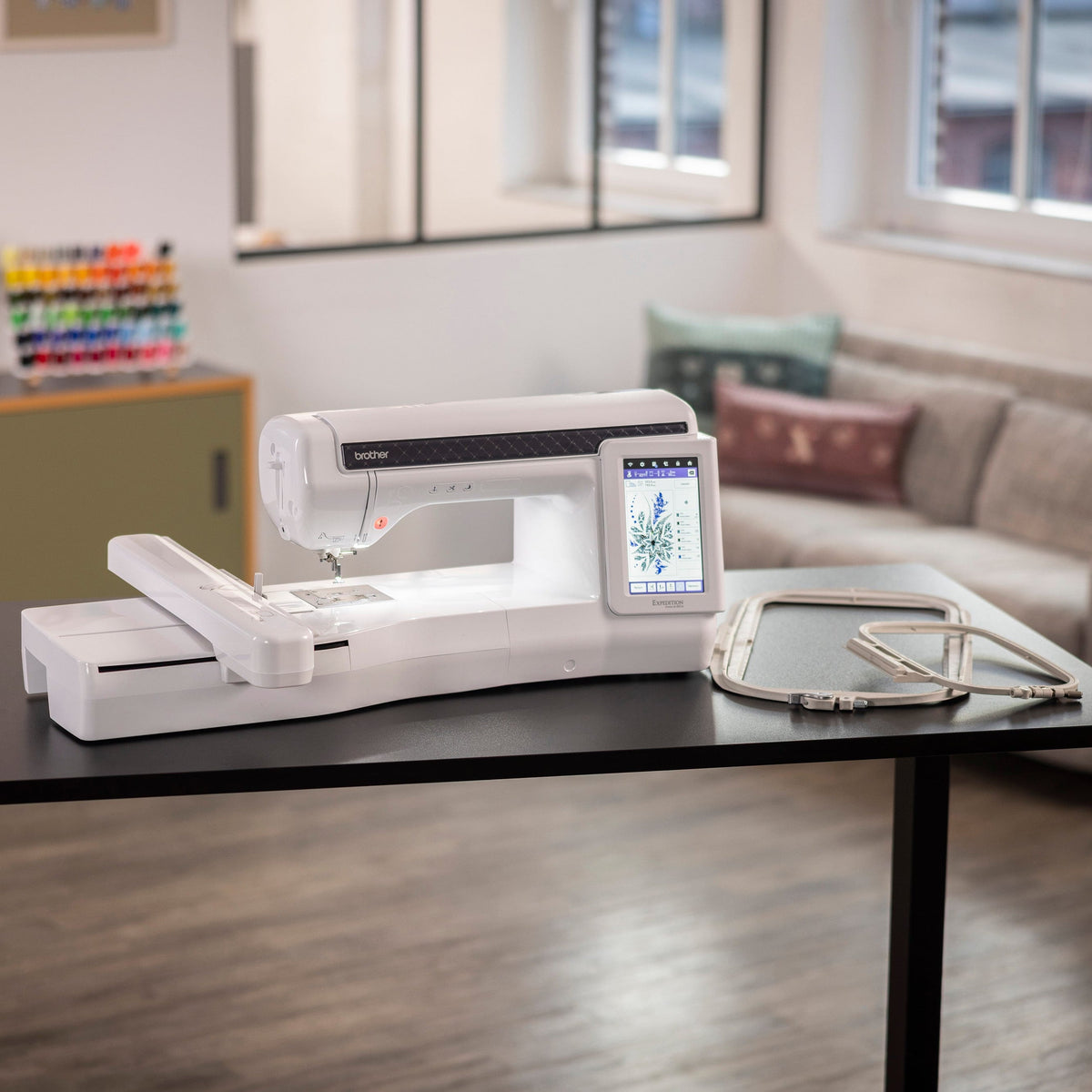 A white sewing machine with an attached embroidery thread spool holder and a small display screen, placed on a black table in a modern living room setting.