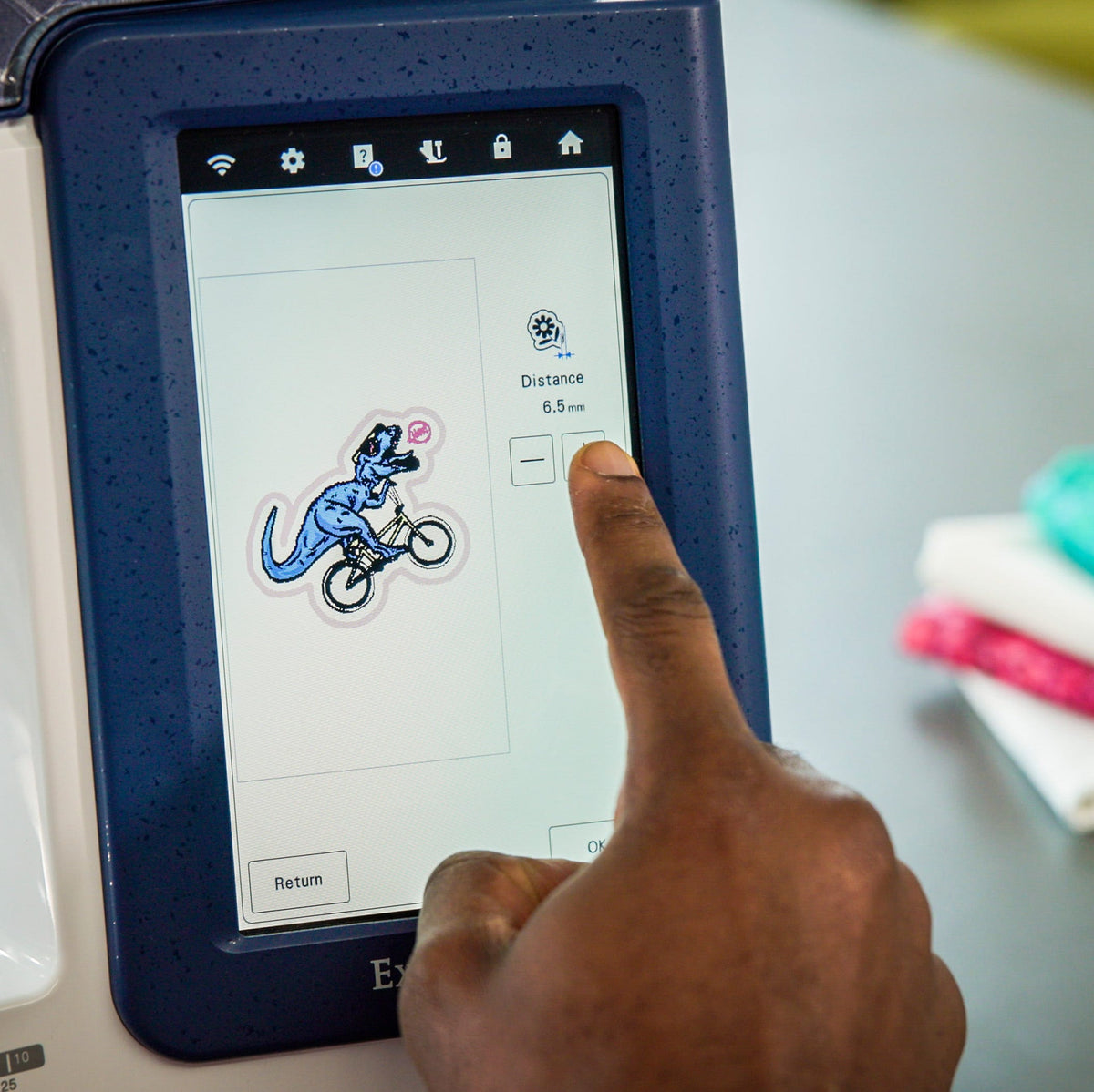 A close-up of a person's finger touching the screen of an embroidery machine, with a blue and white embroidery design visible on the touchscreen. A stack of colorful fabric samples sits on the table in the blurred background.