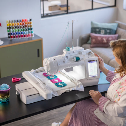 A woman sits at a table using a white embroidery machine in a bright room. The machine has a lit touchscreen and is stitching a colorful floral design on a white fabric.