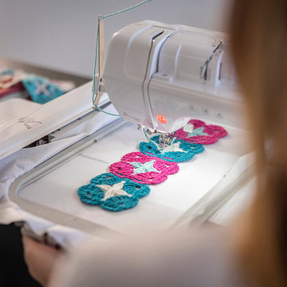 A close-up lifestyle shot of a person using a white sewing machine to embroider four crocheted flower motifs in teal, pink, and white. The person is shown from behind, with their hair blurred in the foreground, and the sewing machine is in sharp focus.