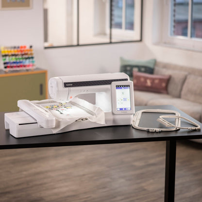 A white Brother embroidery machine with a digital display screen and a white bobbin case, set on a black table in a modern, softly blurred living room environment.