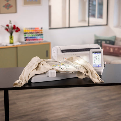 A white embroidery machine sits on a black table in a bright, modern living room. A beige garment is draped over the machine's embroidery bed, and a small stack of fabric samples rests on top.