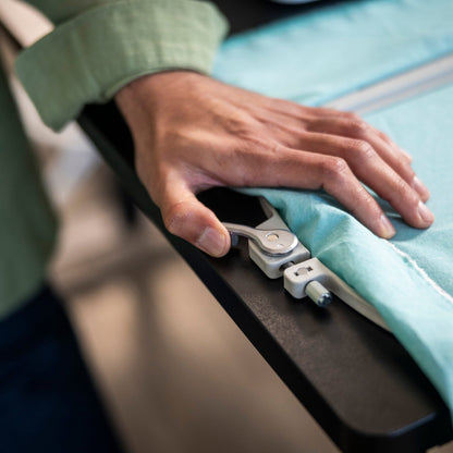 A close-up of a person's hand operating a white and silver electronic fabric measurer on a light blue garment laid out on a black table.
