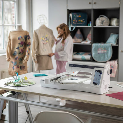 A white digital embroidery machine sits on a light wood table in a bright sewing room, with a woman standing in the background near mannequins and storage shelves.