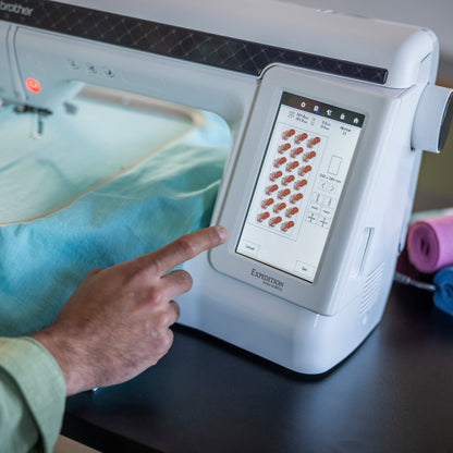 A close-up of a Brother embroidery machine with a person's hand touching the touchscreen interface, with light blue fabric inside the embroidery hoop and rolls of thread on a table.