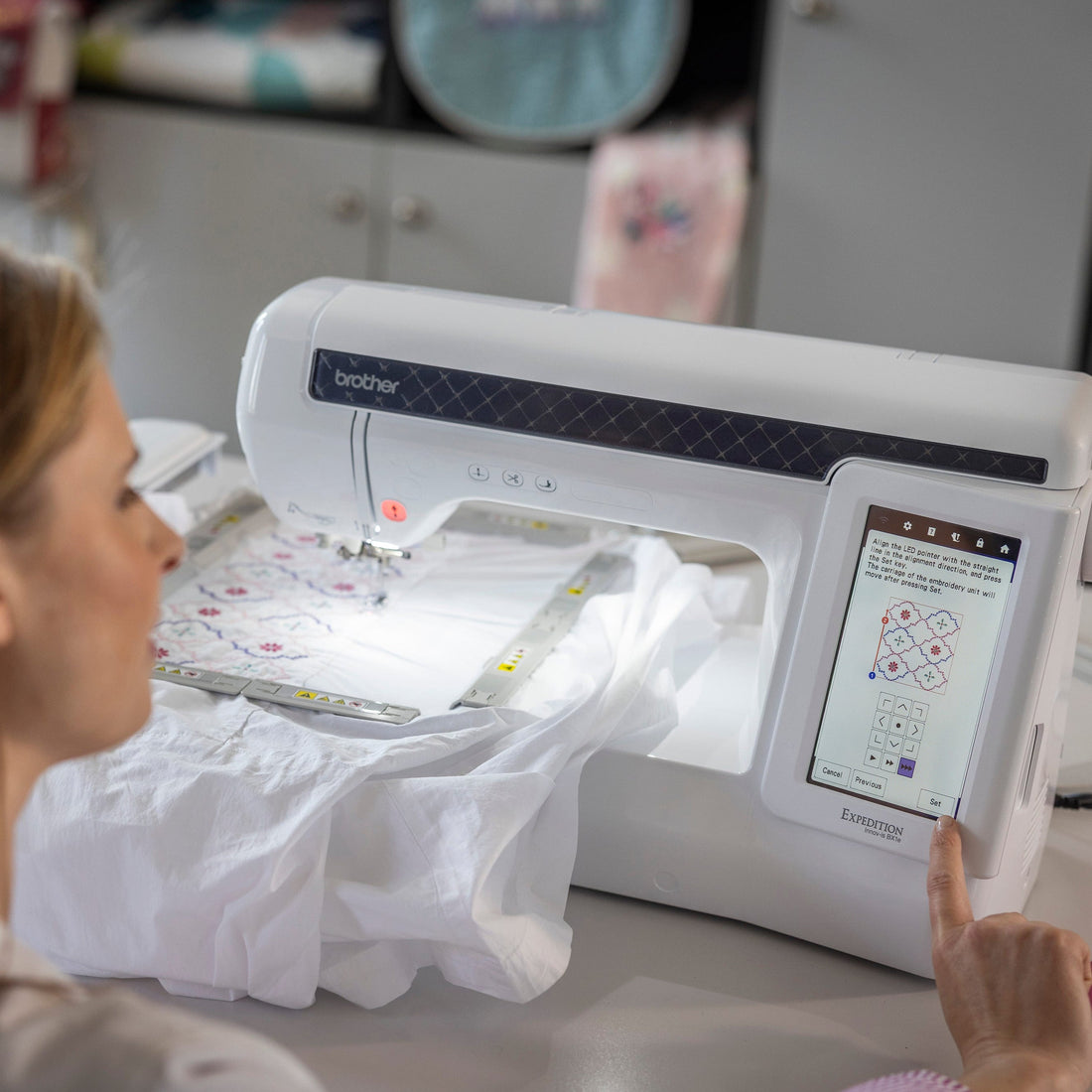 A woman uses a Brother embroidery machine to create a white embroidered garment in a bright, modern craft room.