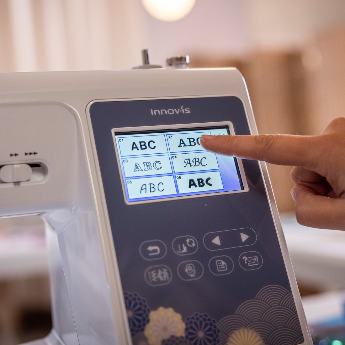 A close-up of a white Innovis sewing machine with a dark control panel and a person's finger touching the touchscreen display, set against a softly blurred indoor background.