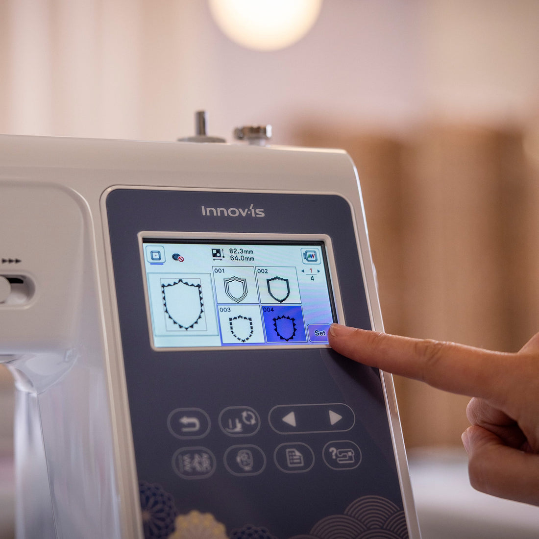 A close-up of a white Innovis sewing machine with a dark gray control panel and a finger touching the touchscreen display, set against a softly blurred indoor background.