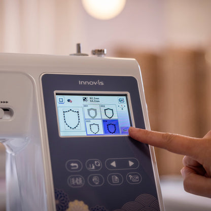 A close-up of a white Innovis sewing machine with a dark gray control panel and a finger touching the touchscreen display, set against a softly blurred indoor background.