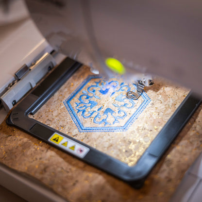 A close-up of an embroidery machine in operation, stitching a blue and white geometric pattern onto a textured beige fabric under warm lighting.
