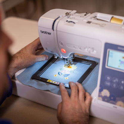 A close-up lifestyle shot of a person using a Brother embroidery machine to embroider a cartoon character onto a square fabric sample. The person is wearing a red beanie and looking down at the machine's needle area.
