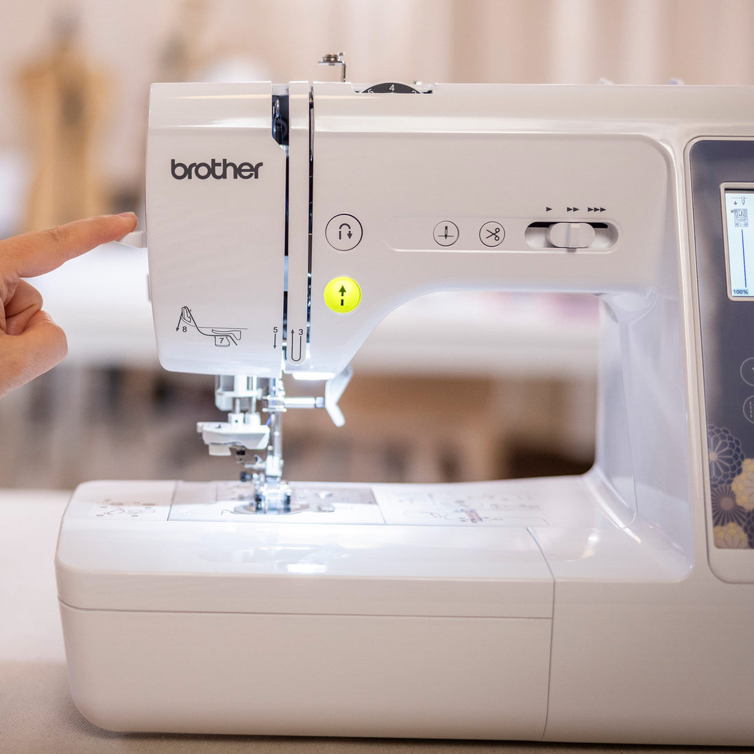 A close-up of a Brother Innov-is sewing machine with a person's hand pointing at the upper left section of the machine, set against a softly blurred indoor background.