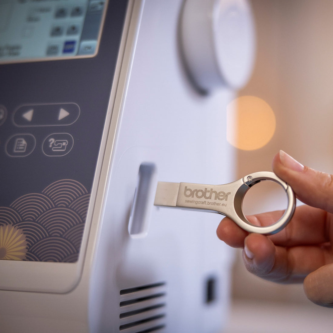 A close-up of a hand holding a Brother USB flash drive with a keyring loop, positioned next to the control panel of a Brother sewing machine.