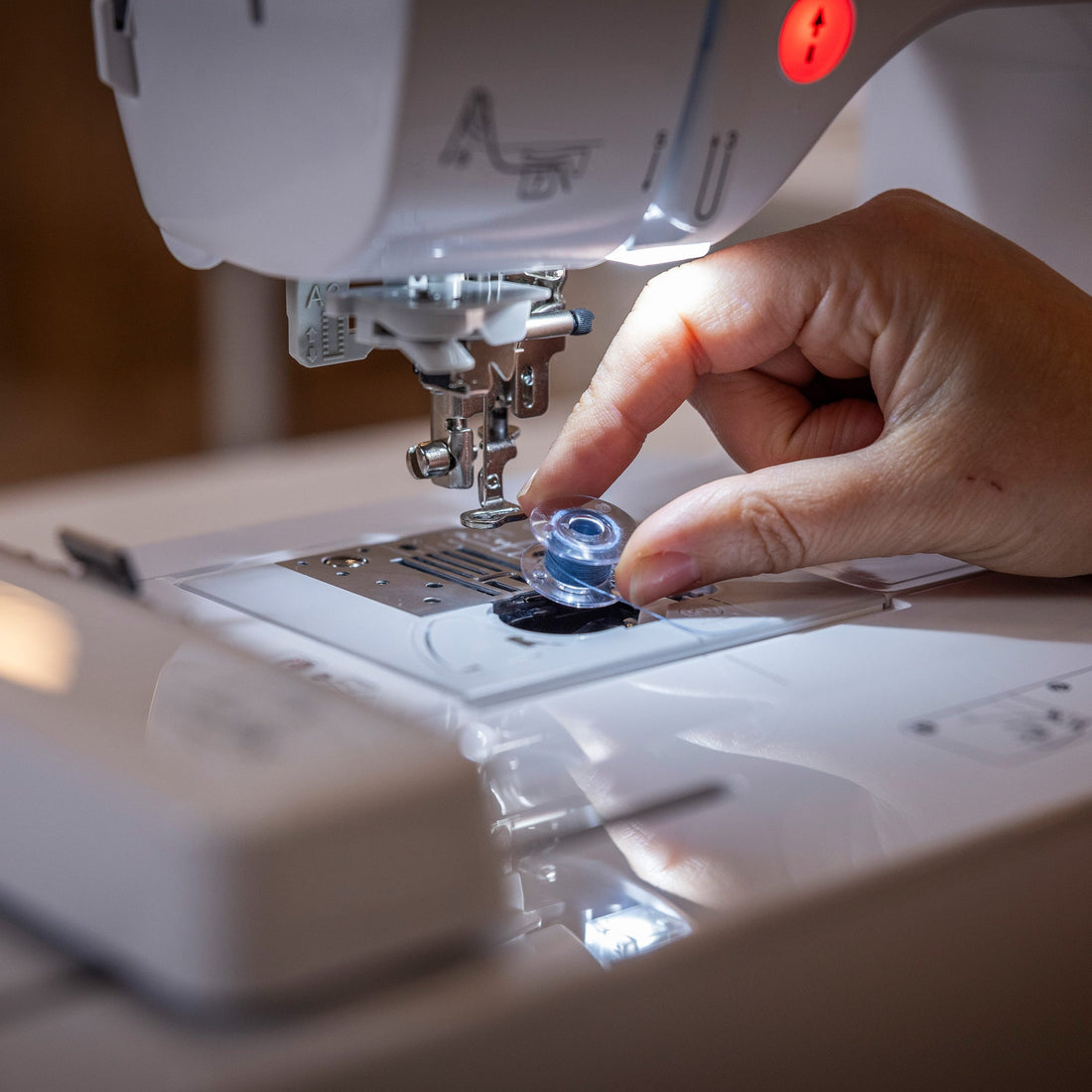A close-up of a person's hand placing a small blue bobbin into the needle area of a white sewing machine under built-in lighting.