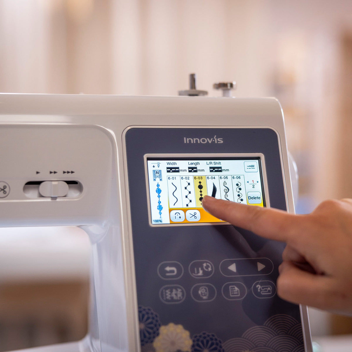 A close-up of a white Innovis sewing machine with a person's finger touching the touchscreen display, set against a softly blurred indoor background.
