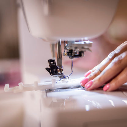 A close-up shot of a person's hand with pink nail polish guiding fabric under the needle of a white sewing machine.