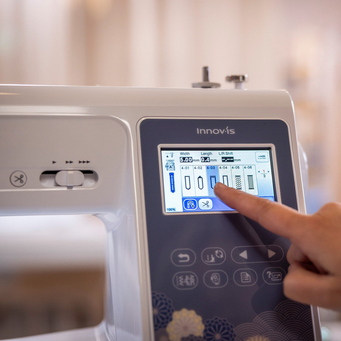 A close-up of a white and navy Innovis sewing machine with a person's finger touching the touchscreen display, set against a softly blurred indoor background.