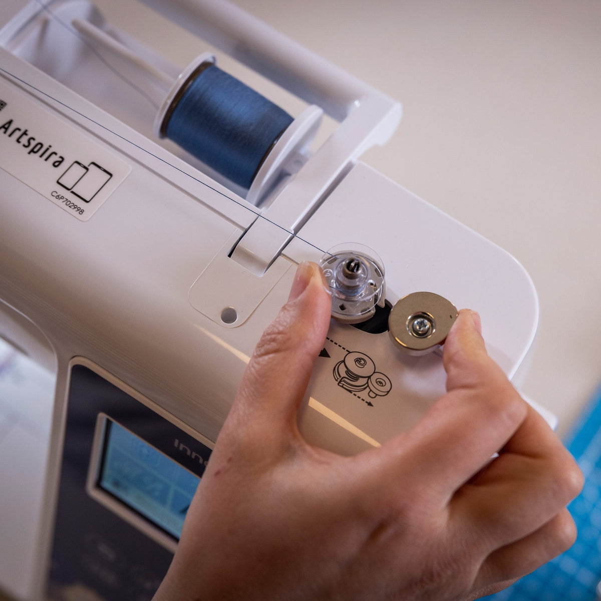 A close-up of a person's hand adjusting the thread spool and tension settings on a white Artspira sewing machine, with a blue cutting mat visible in the background.