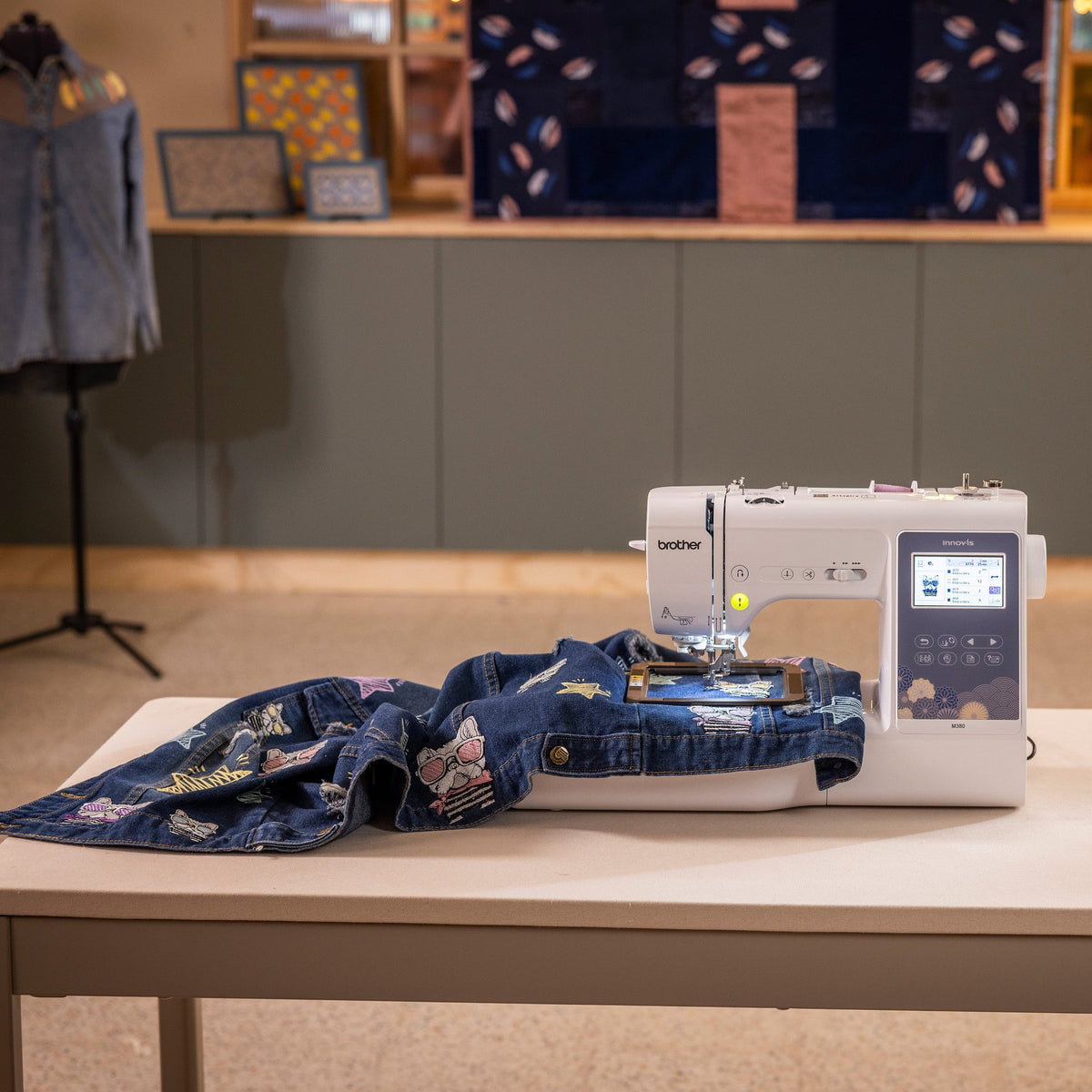 A Brother sewing machine sits on a white table in a styled indoor setting, with a blue denim garment laid out on the machine bed. The background features a gray cabinet, a mannequin wearing a denim shirt, and warm ambient lighting.