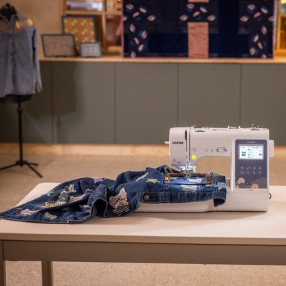 A Brother sewing machine sits on a white table in a styled indoor setting, with a blue denim garment laid out on the machine bed. The background features a gray cabinet, a mannequin wearing a denim shirt, and warm ambient lighting.