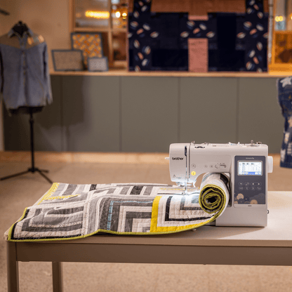 A Brother sewing machine sits on a white table in a sewing studio, with a patterned fabric roll under the needle. In the background, mannequins display a blue star-patterned vest and a blue apron.