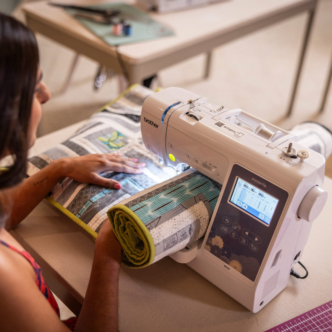A woman using a white Brother sewing machine with a digital LCD screen, stitching a rolled fabric sample on a table in a bright room.