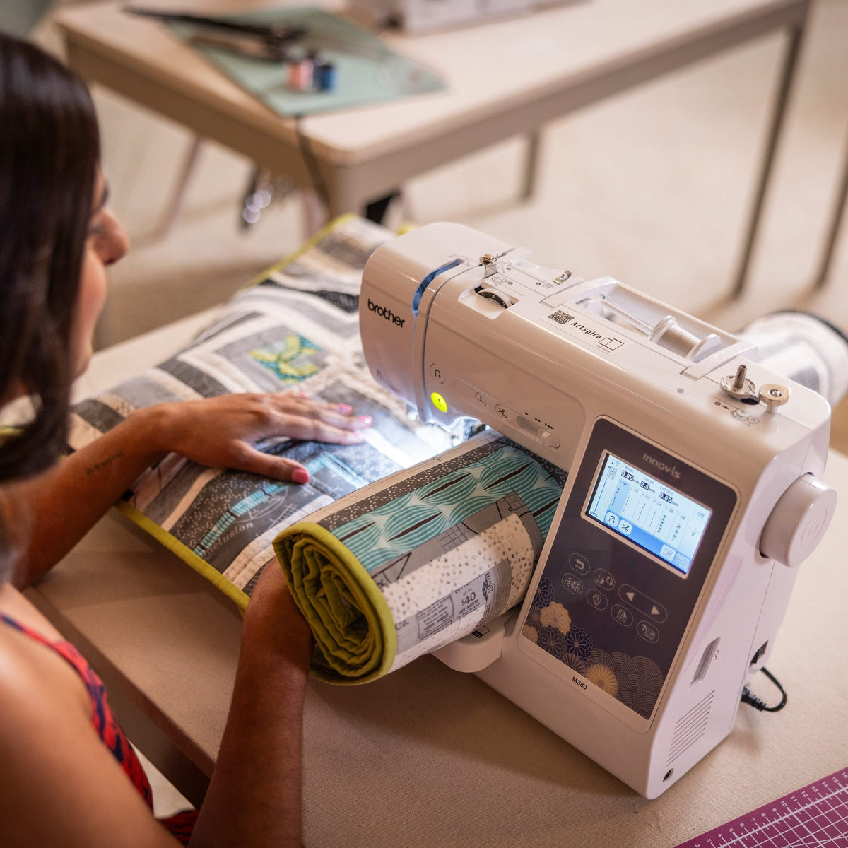 A woman using a white Brother sewing machine with a digital LCD screen, stitching a rolled fabric sample on a table in a bright room.
