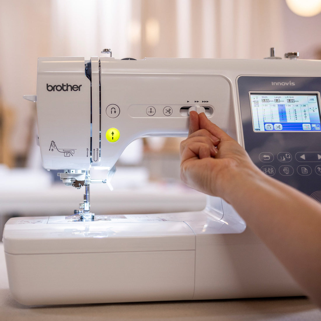 A Brother sewing machine with a digital touchscreen interface, shown in a bright, softly blurred indoor setting. A person's hand is reaching toward the control panel.