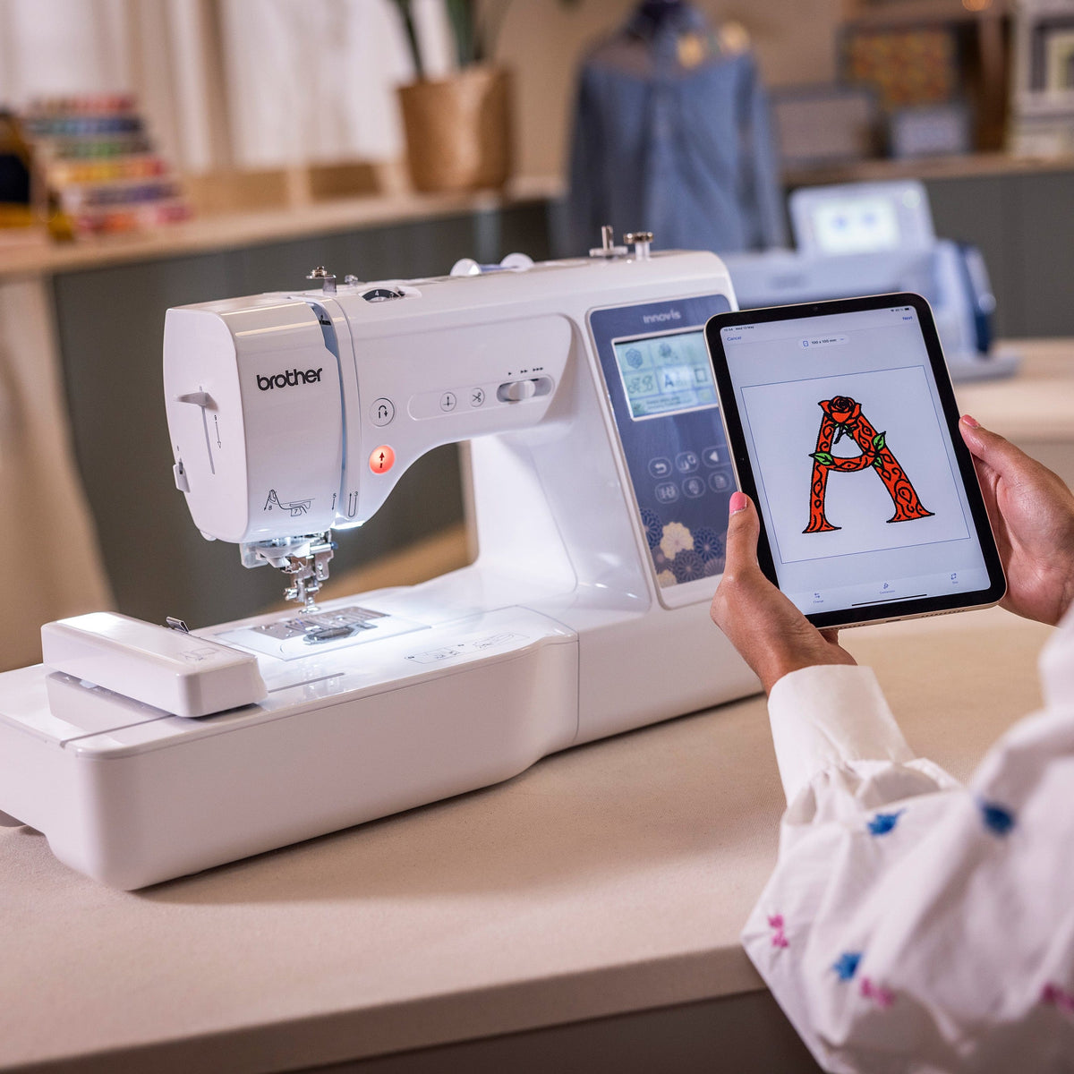 A person holds a tablet displaying a red letter 'A' pattern while using a white Brother sewing machine in a bright, modern sewing room.
