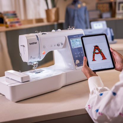 A person holds a tablet displaying a red letter 'A' pattern while using a white Brother sewing machine in a bright, modern sewing room.