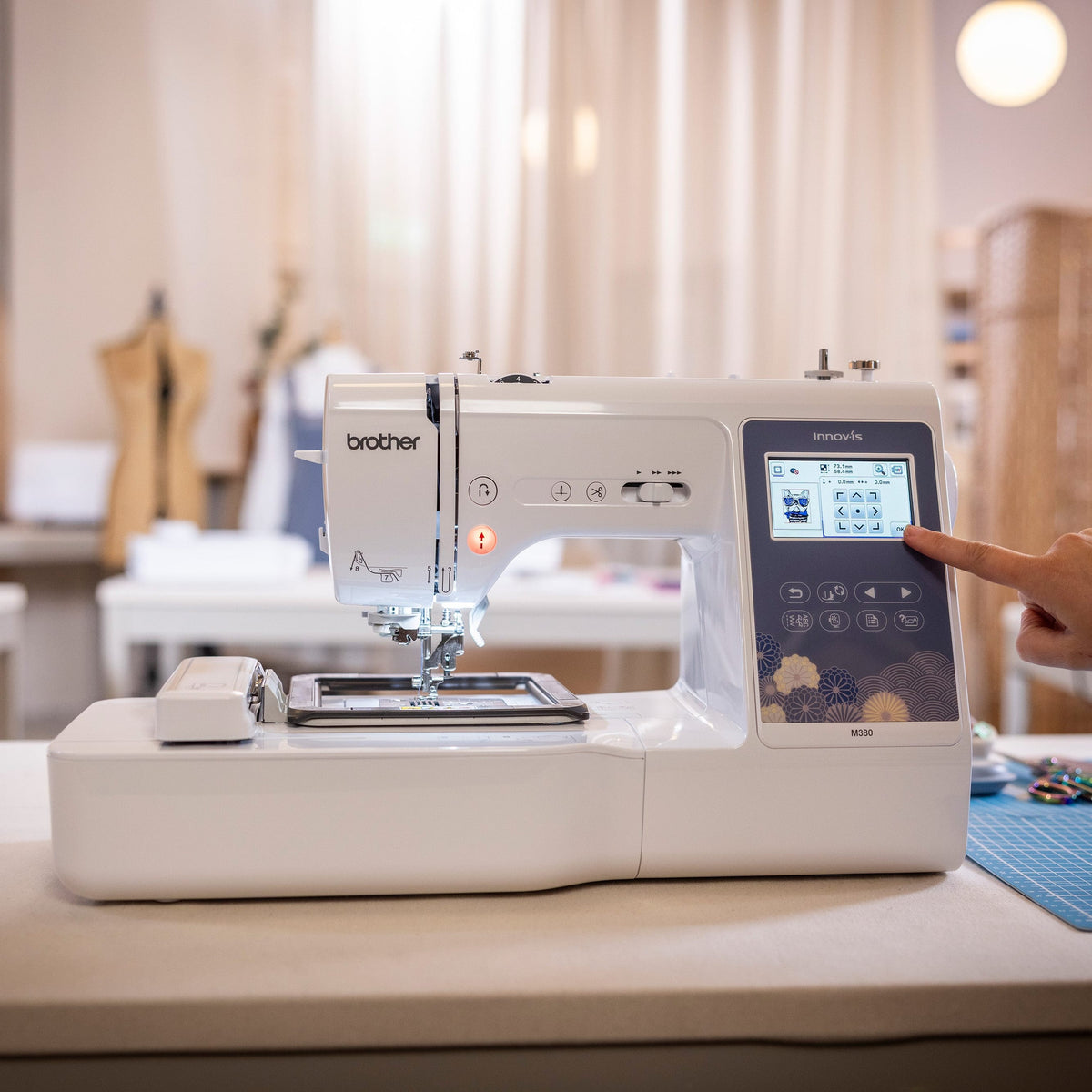 A Brother sewing machine on a table in a softly lit craft room, with a person's hand pointing at the digital touch screen display.