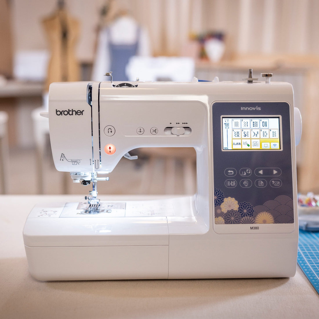 A Brother Innov-is sewing machine in white and dark gray, placed on a table in a softly blurred sewing room with a blue cutting mat nearby.