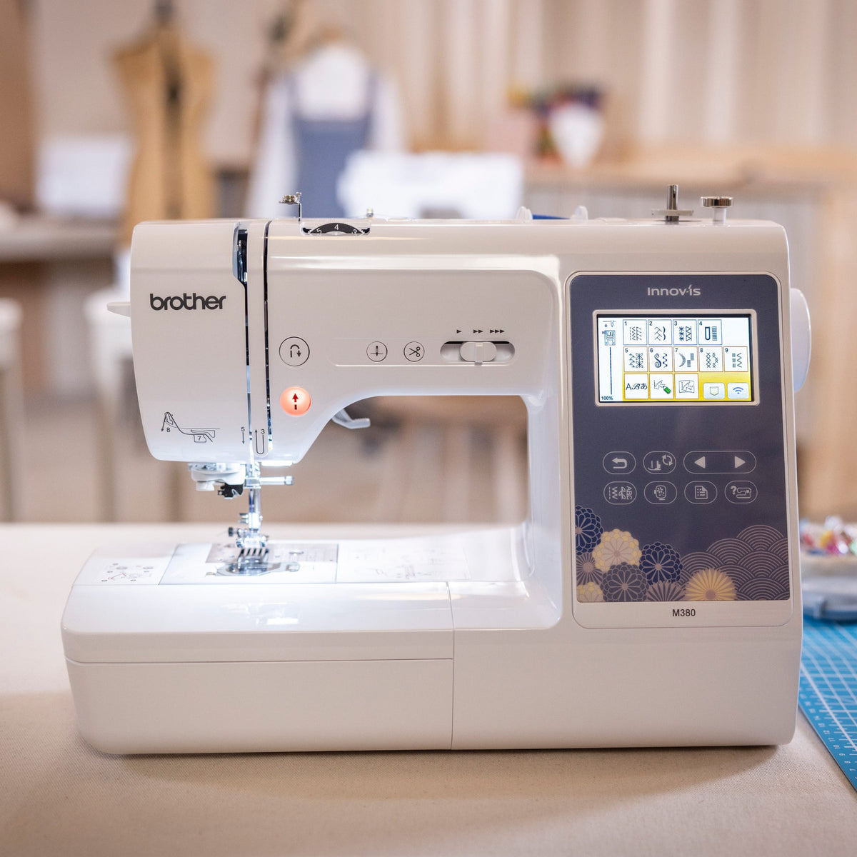 A Brother Innov-is sewing machine in white and dark gray, placed on a table in a softly blurred sewing room with a blue cutting mat nearby.