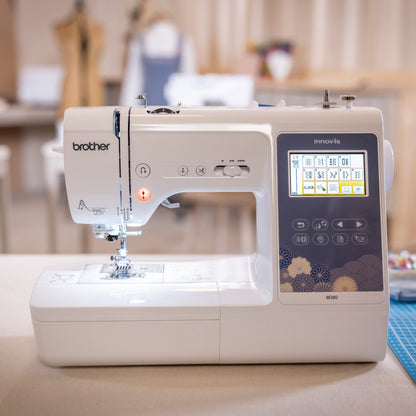 A Brother Innov-is sewing machine in white and dark gray, placed on a table in a softly blurred sewing room with a blue cutting mat nearby.