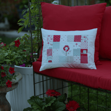 A square decorative throw pillow with a red and white patchwork holiday design, resting on a red outdoor sofa cushion in a garden setting.