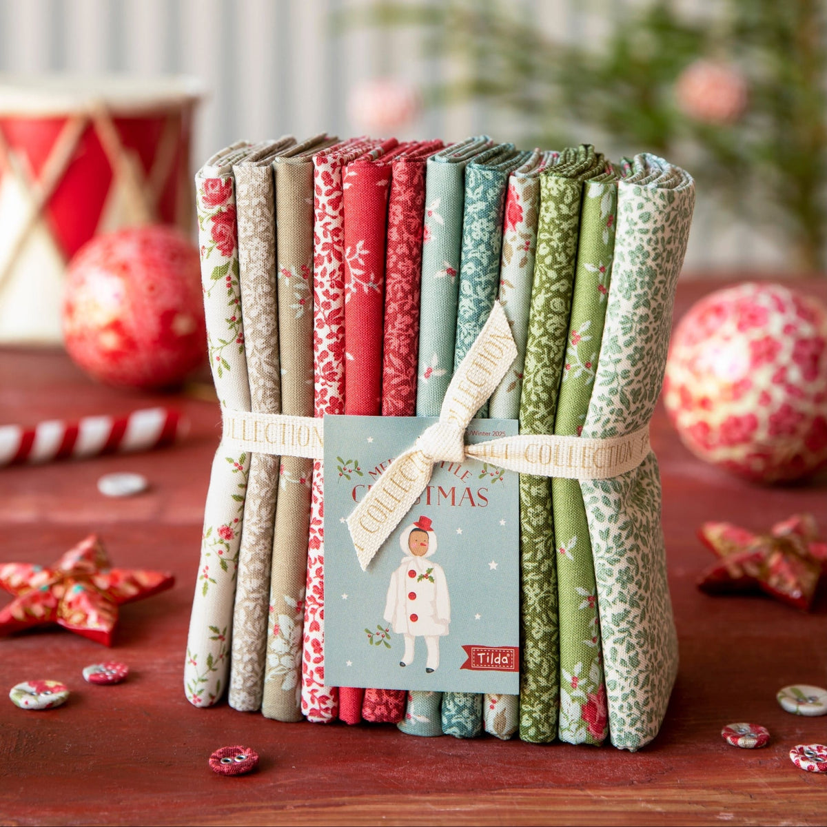 A bundle of folded quilts tied with a ribbon and card, displayed on a wooden surface with holiday decorations in the background. The quilts feature various festive patterns in red, green, blue, and beige.