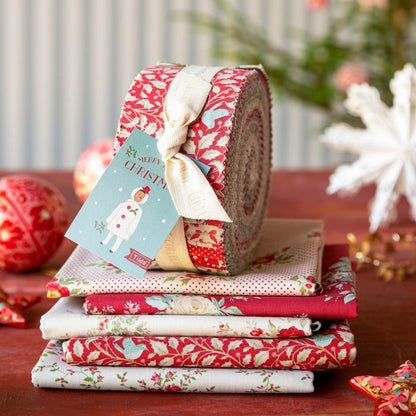 A stack of folded and rolled fabric bundles wrapped in red holiday patterns, tied with a cream ribbon, and labeled with a blue 'Cozy Christmas' tag. The fabrics rest on a wooden surface with blurred festive decorations in the background.